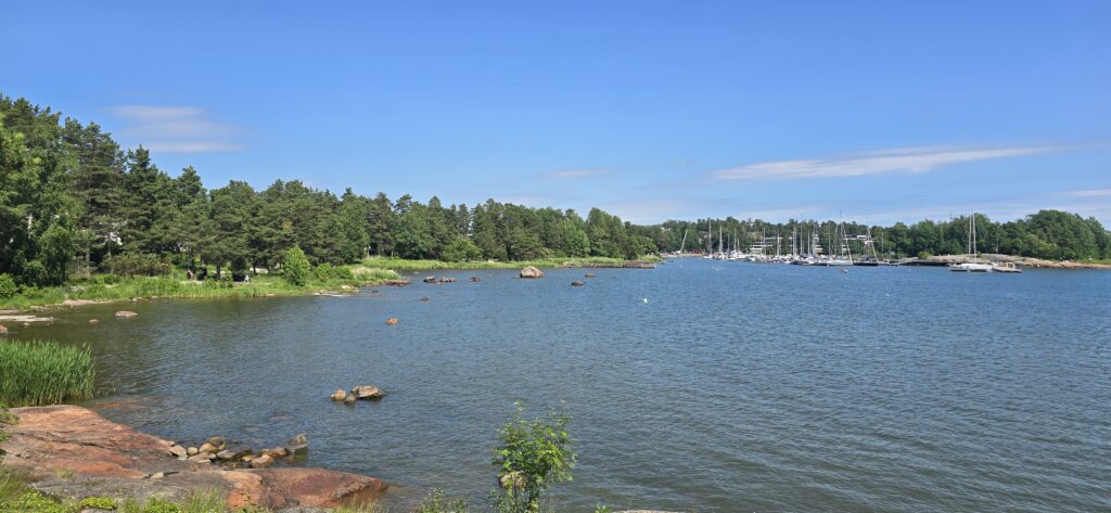 Aurinkoinen pohjoismainen merimaisema Espoon rantaraitilta: kallioista rantaa, havumetsää ja pieni venesatama purjeveneineen kirkkaan sinisen taivaan alla.